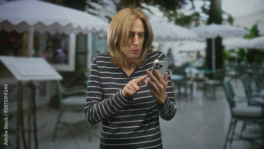 Fototapeta premium Woman tapping smartphone with visible hands and furrowed expression at a street cafe terrace, holding phone while scrolling through messages; everyday tech frustration.