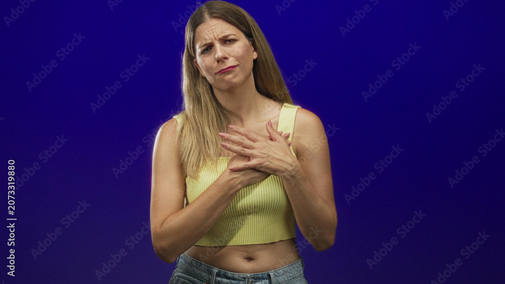 Fototapeta premium Young woman clutching chest with hands in a purple studio, wearing yellow tanktop and jeans; pain anxiety.