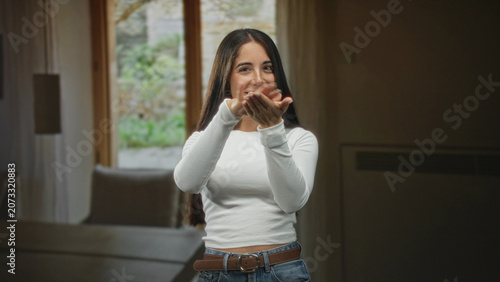 Obraz na plátně Young woman smiling beckons with hands in a sunlit building living room by a window and sofa; warm welcome joy