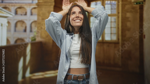 Foto Young woman lifts hands above head showing palms, smiling with long hair, wearing white crop top, open blue shirt, jeans and belt in a sunlit building corridor; joyful
