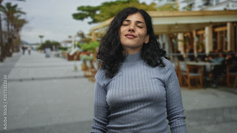 Fototapeta premium Young brunette woman standing with closed eyes and slight smile on street beside a restaurant terrace outdoors; calm reflection.