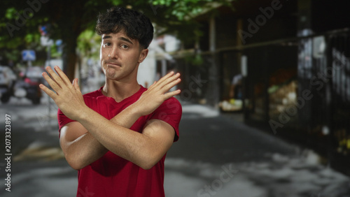 Konstfotografi Young man with bare forearms arms crossed x on a city street wearing red t shirt facing camera, palms visible; rejection doubt