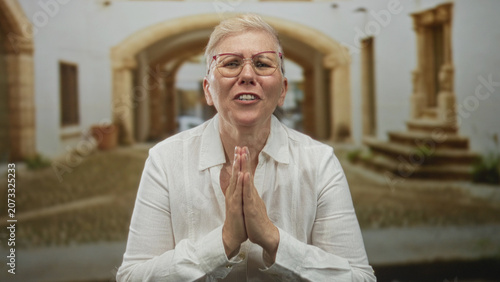 Quadro su tela Woman with hands pressed together at chin on an old town street wearing glasses and white shirt in front of archway; hope gratitude devotion