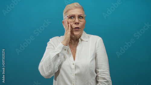 Foto Woman with hand to mouth and open mouth gesture wearing glasses and white shirt in teal studio; surprise reaction moment