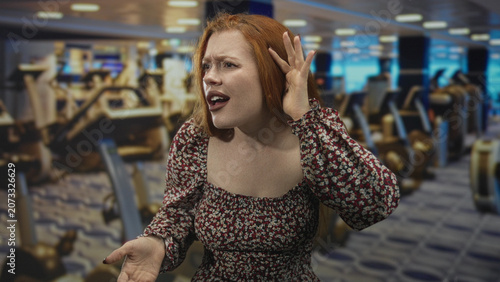 Canvastavla Redhead woman cups ear and points finger toward camera amid gym cardio machines in building, leaning forward with puzzled frown and raised hand; frustration confrontation