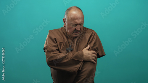 Obraz na plátně Man in brown robe with cross necklace clutches his chest and upper arm in pain in studio; suffering prayerful