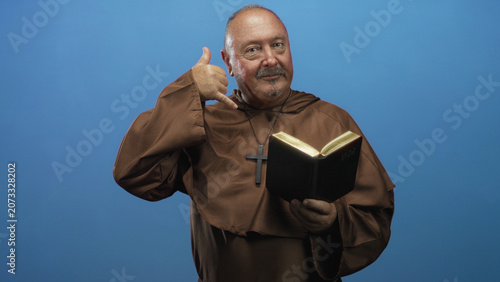 Photos Man monk in brown robe holds bible and makes phone gesture with hand to ear in studio; playful devotion