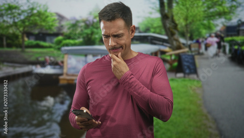 Fotografie Man holding smartphone and touching chin on canal towpath near a moored boat and bridge, reading screen with a furrowed brow and thoughtful expression; pensive