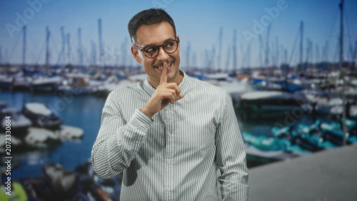 Cuadro en lienzo Hispanic man with moustache and glasses, finger to lips shushing at marina with boats and masts in view; playful secrecy