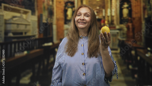 Obraz na plátně Senior woman holding lemon in church pew, striped blouse, smiling with eyes closed amid ornate wooden altar; serenity gratitude