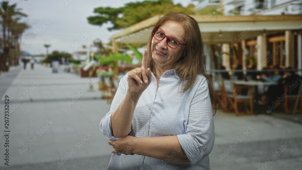 Fototapeta premium Woman points finger up at chest level on a street beside an outdoor cafe, wearing red glasses and a striped shirt; gentle advice warmth.