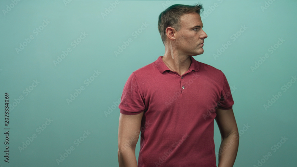 Fototapeta premium Man in red polo scratches neck with hand behind neck in studio teal backdrop, visible forearm and pensive expression; thoughtful uncertainty.
