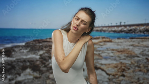 Fotografie Woman holding neck and shoulder with hand on rocky beach, wincing in discomfort as wind tugs hair; pain vulnerability
