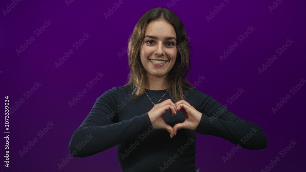 Obraz premium Young woman smiling and forming a heart with both hands in studio with deep purple backdrop; love kindness.