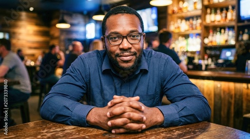 Man in blue shirt with glasses sitting at a table in a bar.