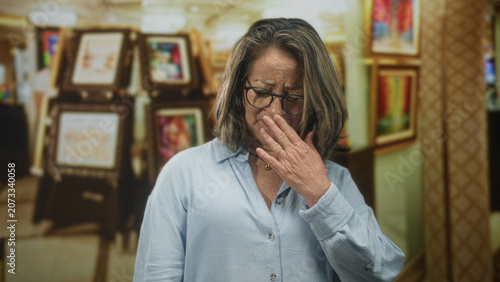 Fotografía Hispanic woman with grey hair pinches nose with hand amid framed art in building; disgust odor concern