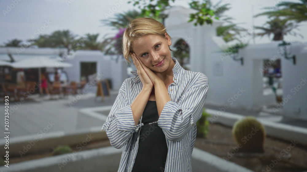 Fototapeta premium Caucasian blond woman presses hands to cheek, eyes closed in a gentle resting pose in sunlit street courtyard with cafes; serenity.