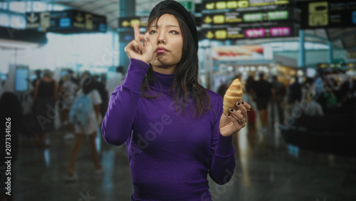 Tableau sur toile Woman in purple sweater holds croissant and pinches crumb with fingers while glancing at departure boards inside an airport terminal; contemplation travel
