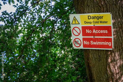 Danger sign warning of deep cold water. No access. No swimming. Warning sign on a tree with trees and lake behind.