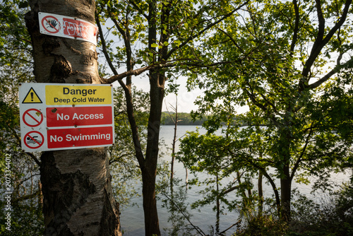 Danger sign warning of deep cold water. No access. No swimming. Warning sign on a tree with trees and lake behind.
