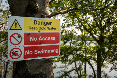 Danger sign warning of deep cold water. No access. No swimming. Warning sign on a tree with trees and lake behind.