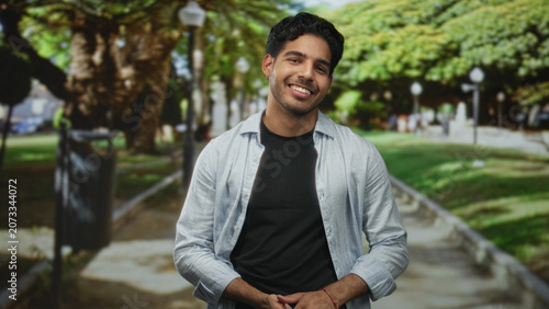 Photos Man smiling with hands clasped at waist on a tree lined street path, wearing light shirt over black tee; warmth contentment