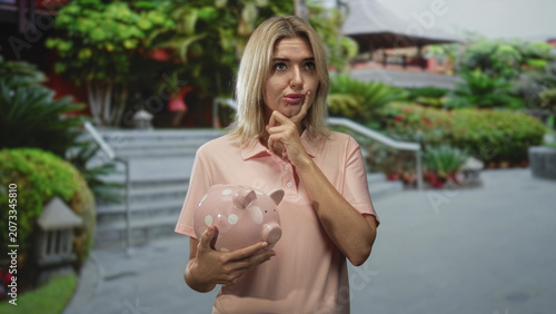 Obraz na płótnie Woman young blonde holds pink polka dot piggy bank, finger to chin in front of building steps and landscaped courtyard, pensive expression; saving thoughtful