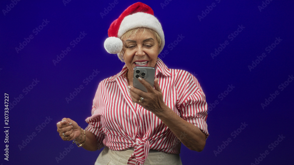 Fototapeta premium Woman senior hispanic in red striped shirt and santa hat taps smartphone with both hands and looks at screen in studio; holiday cheer.