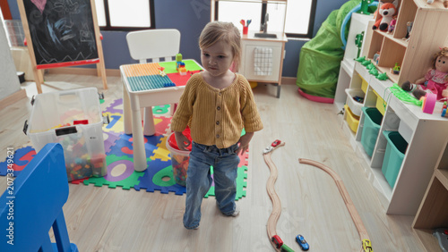 Tableau sur toile Girl standing with toy train on foam playmat in playroom with shelves and chalkboard; quiet curiosity