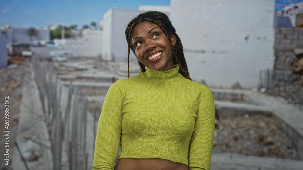 Fototapeta premium Woman smiling, showing teeth and looking up at a construction site, wearing lime green crop top with exposed midriff and braided hair; playful confidence.