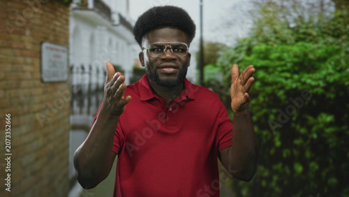Fotografie Man gesturing with hands on street beside brick building and hedgerow, wearing red polo and glasses; persuasive discussion