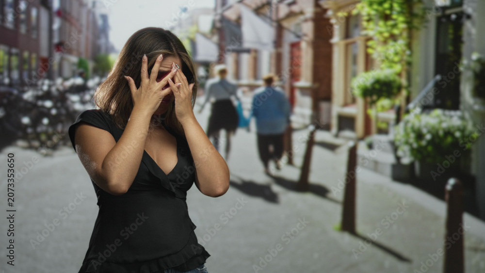 Fototapeta premium Woman covering mouth with hands on street, wearing black blouse and glasses near bollards and storefronts; shock vulnerability.