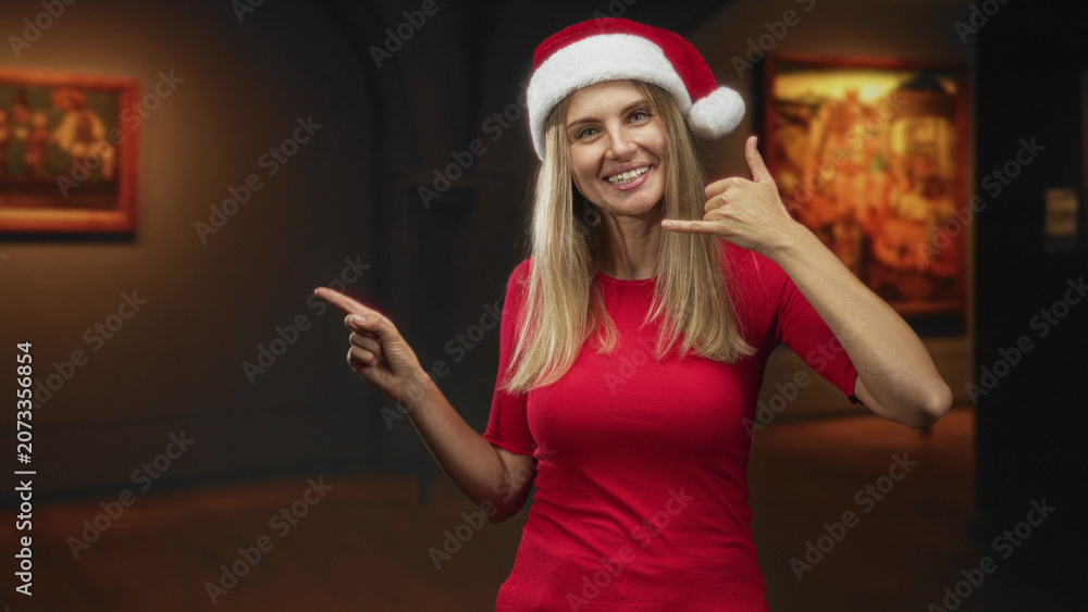 Fototapeta premium Woman in santa hat points left with index finger and makes call gesture with right hand inside a dim museum building; festive joy.