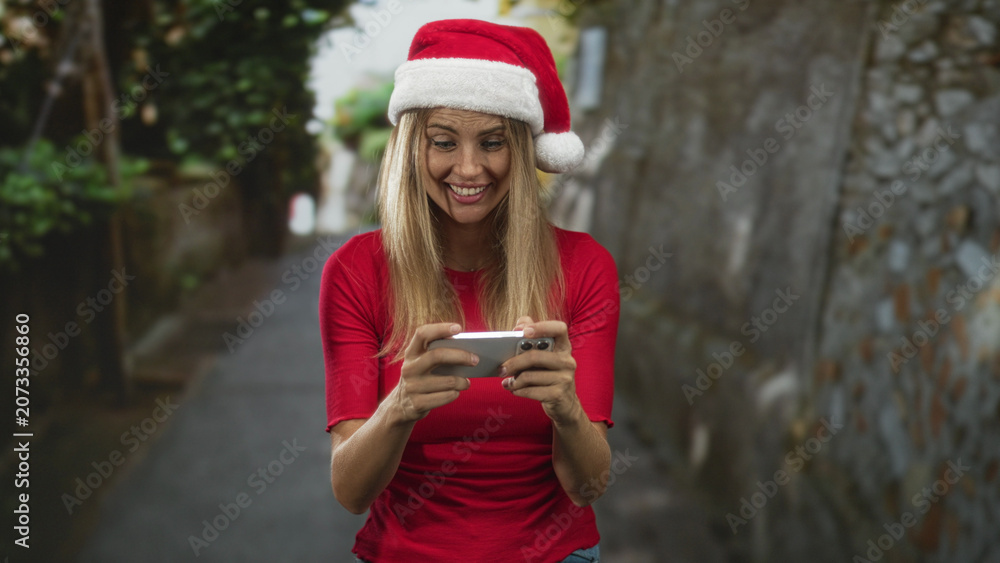 Fototapeta premium Blonde woman holding smartphone with both hands, smiling and looking down while wearing a santa hat on a narrow street; festive joy.