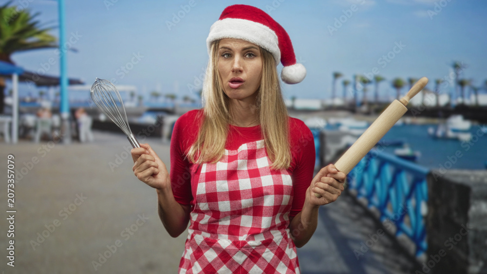 Fototapeta premium Woman in santa hat and red check apron, hands holding whisk and rolling pin on street near a marina pier with palm trees and boats; holiday fatigue.