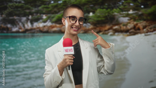 Photos Woman holding red microphone labeled news points index finger to cheek while smiling at camera on a sandy beach cove wearing glasses white blazer black crop top; broadcast confidence