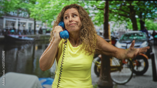Obraz na płótnie Middle aged hispanic woman holds blue telephone handset to ear, hand on hip and rolling eyes in a street scene by a canal with parked bikes; exasperation