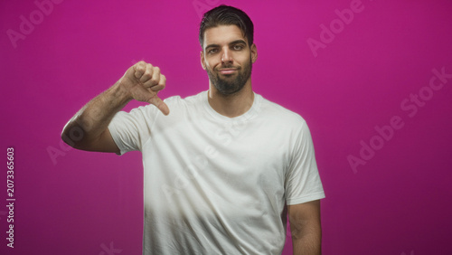 Bild auf Leinwand Hispanic man with beard wearing a white t shirt giving thumb down gesture with exposed hand and forearm against a magenta studio backdrop, slight smirk visible; disapproval rejection