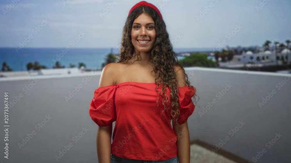 Fototapeta premium Teenage hispanic woman with bare shoulders in a red off shoulder top smiling and looking up on a rooftop building terrace by the sea; carefree joy.