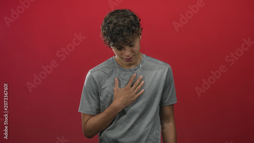 Fotografie Teenage man places hand on chest with eyes closed and calm expression against red studio backdrop; sincerity gratitude