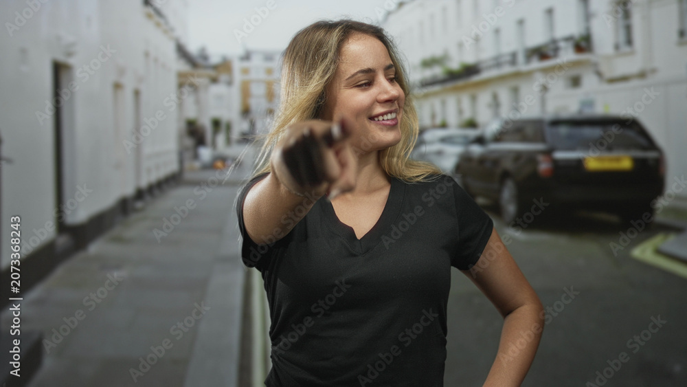 Fototapeta premium Woman pointing finger at camera on narrow city street, smiling and leaning forward, wearing black tshirt; confidence.