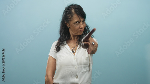 Obraz na plátně Woman senior points index finger to camera in studio wearing white blouse and pendant showing scolding gesture; warning