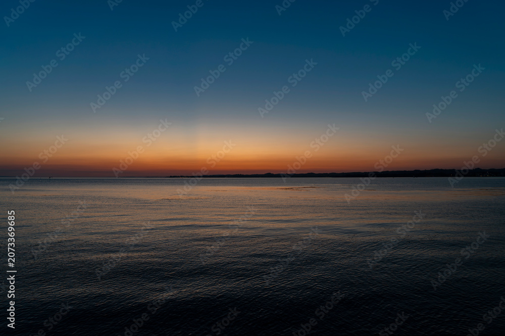 Fototapeta premium Serene panoramic view of Lake Constance after sunset, featuring a clear twilight sky with a vibrant orange glow on the horizon fading into deep blue