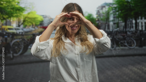 Fotografie Woman smiling and forming heart with hands on a street canalside bridge, wearing white long sleeve shirt; love kindness