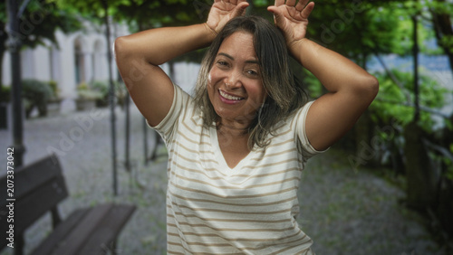 Fotografía Hispanic woman with hands raised making antlers gesture and broad smile on a street bench walkway; joy playful moment