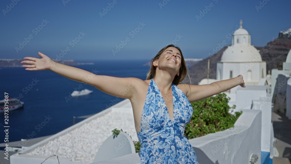Fototapeta premium Woman smiling with arms outstretched on a white church building terrace in santorini overlooking blue sea and island vistas; serenity escape.