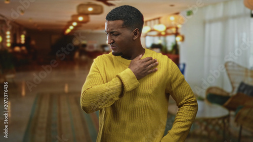 Schilderij op canvas Man in yellow sweater clutching his shoulder with hand and wincing while standing in a hotel lobby reception area; pain health concern