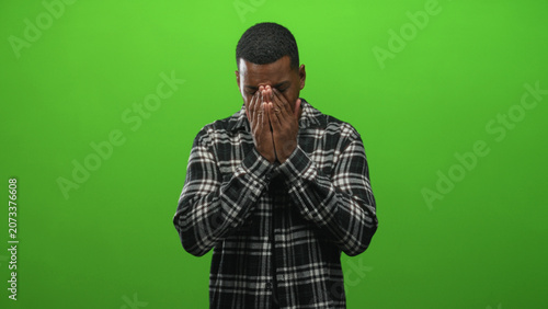 фотографія Young man covering face with hands in green studio with plaid flannel shirt and closed eyes; sadness vulnerability