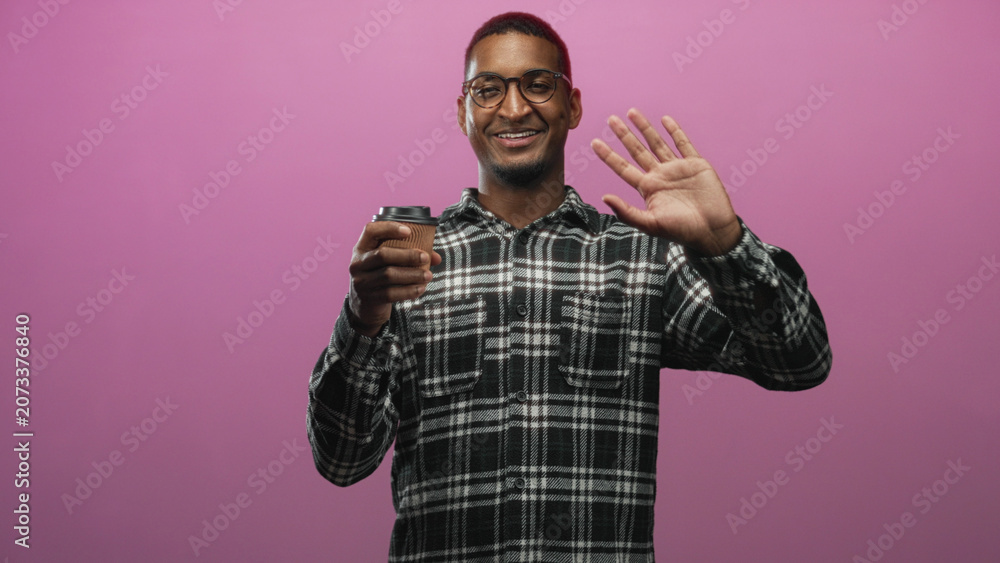 Naklejka premium Man wearing glasses and plaid shirt holding paper coffee cup and waving hand in studio with pink backdrop; friendly greeting.