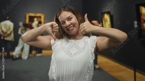 Vászonkép Woman with bare arms showing thumbs up and thumbs down gesture in gallery building; ambivalence choice